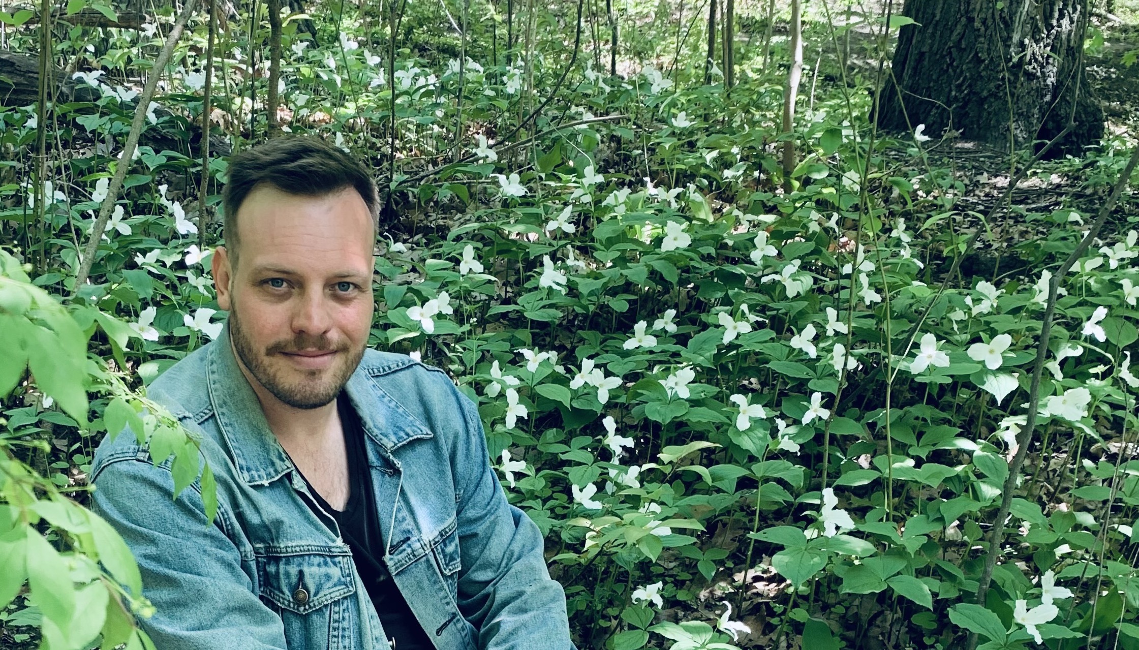 Portrait of Stefan Herda sitting in a forest clearing surrounded by white trillium plants.